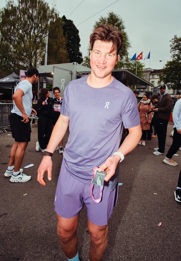 Man in purple athletic wear holding a race medal, standing outdoors with other people and event tents in the background.