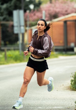 Joven mujer corriendo al aire libre por un camino pavimentado, lleva una chaqueta morada, pantalones cortos negros, calcetines blancos y zapatos deportivos grises.
