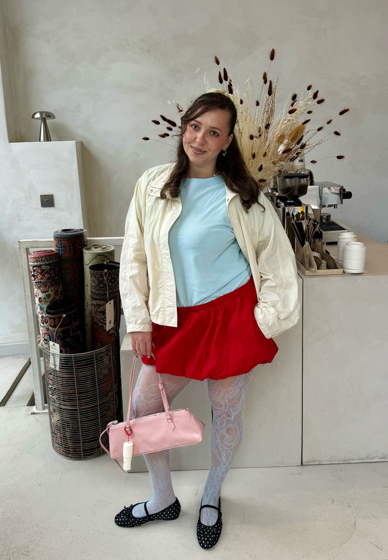 Femme aux longs cheveux bruns debout à l'intérieur portant une jupe rouge, un haut bleu, une veste blanche, des collants à motifs, des chaussures noires et tenant un sac à main rose.