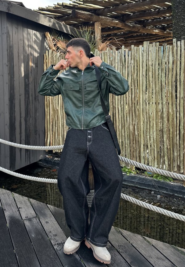 Vīrietis zaļā ādas jaku regulē apkakli, stāvot uz koka klāja pie bambusa žoga un virvju margām virs ūdens.