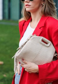 Μπεζ υφασμάτινη τσάντα σε ορθογώνιο σχήμα, με υφή και φούντες, κρατημένη μπροστά από �ένα φωτεινό κόκκινο σακάκι.