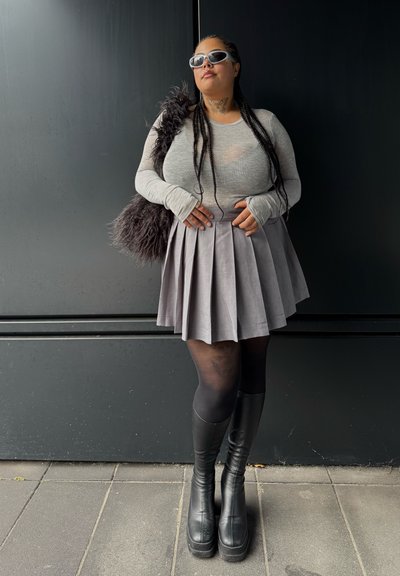 Camiseta gris de manga larga con agujeros para los pulgares, falda gris plisada, botas negras de plataforma hasta la rodilla y gafas de sol blancas de gran tamaño. Bolso de mano negro texturizado.