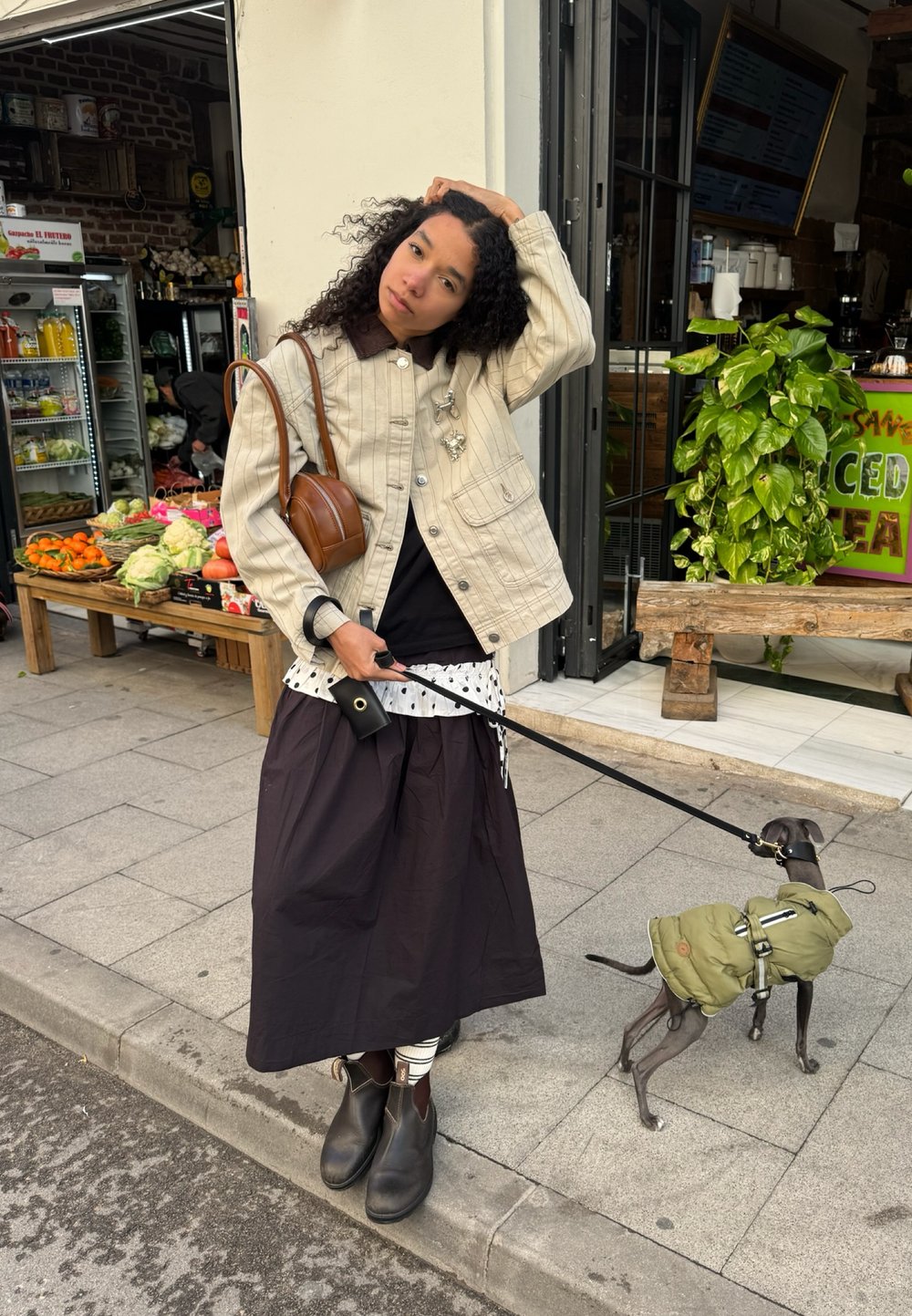 Femme en veste beige et longue jupe sombre se tient sur le trottoir en tenant la laisse d'un petit chien portant une veste verte près d'un magasin de produits frais.