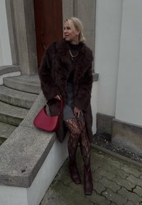 Manteau en fausse fourrure marron, haut noir, jupe grise, collants à motif de dentelle, sac à main bordeaux et bottines à talons assorties.