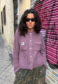 Femme aux cheveux bouclés et foncés portant des lunettes de soleil noires, une veste en tricot violette et un pantalon camouflage, debout devant un volet métallique couvert de graffitis violets.
