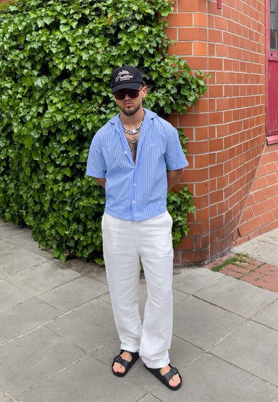 Hombre con camisa de manga corta a rayas azules, pantalones blancos, sandalias negras, gafas de sol y gorra negra, de pie junto a una pared de ladrillo con hiedra.