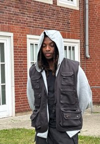 Gilet utilitario nero con più tasche indossato sopra un maglione con cappuccio grigio. Sullo sfondo si vede un edificio in mattoni rossi e il pavimento.