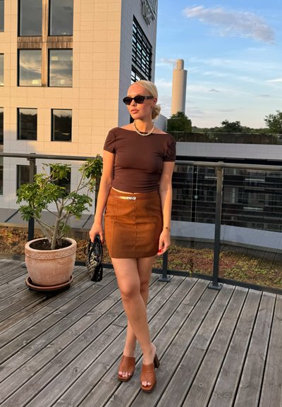 Top marrón ajustado, falda mini de ante color bronce, tacones de plataforma marrones. Bolso negro y gafas de sol. Entorno al aire libre con plantas y una terraza de madera.