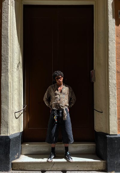 Camisa de botones de estampado de leopardo, pantalones cortos amplios negros, llavero colorido, zapatillas oscuras, de pie en escalones de mármol cerca de puertas marrones.
