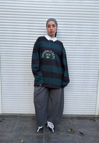 Jeune personne portant une chemise rayée surdimensionnée, un pantalon large gris, un foulard et des chaussures noir et blanc, debout devant une porte à volets blanche.