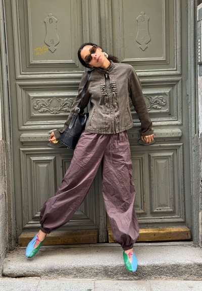 Mujer con gafas de sol, chaqueta marrón suelta, pantalones morados anchos y zapatos coloridos se recuesta contra una puerta doble gris ornamentada al aire libre.
