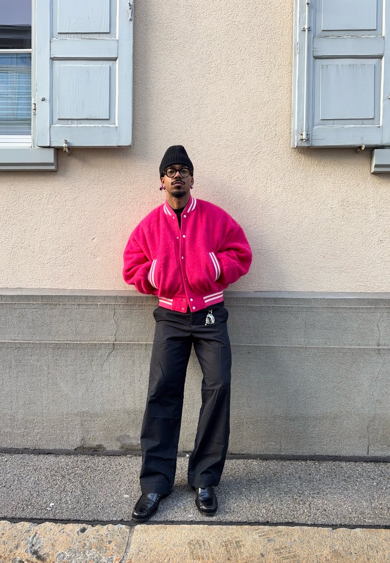 Man wearing bright pink jacket, black beanie, glasses, and black trousers stands with hands in pockets against beige wall with two closed blue shutters.
