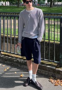 Mlađi muškarac u naočalama za sunce i prugastom puloveru stoji pored ograde u parku, drži zeleni napitak sa slamkom i sluša slušalice.