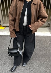 Hnědá semišová bunda přes černý kardigan, v kombinaci s černými širokými kalhotami a sladěnou černou koženou taškou a botami. Texturovaný, ležérní vzhled.