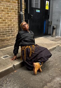 Svart bomberjakke, lang svart skjørt med gule detaljer ved midjen, og brune støvler. Plassert foran en murvegg og en dør.