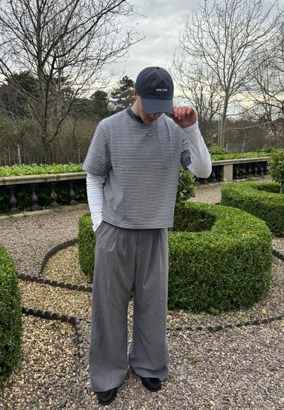 Top de manga corta a rayas combinado con pantalones sueltos grises con rayas. Gorra negra con texto y zapatos negros. Fondo de jardín natural.