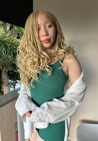 Πράσινη υφασμάτινη ολόσωμη φόρμα συνδυασμένη με ένα χαλαρό λευκό πουκάμισο με κουμπιά; μακριά, σγουρά, ξανθά μαλλιά; ανοιχτόχρωμο δέρμα; εσωτερικός χώρος με ορατή βλάστηση.