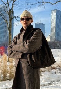 Homme portant un bonnet beige, des lunettes de soleil, un manteau à carreaux et portant un grand sac sombre, debout en plein air dans la neige avec des gratte-ciel modernes en arrière-plan.