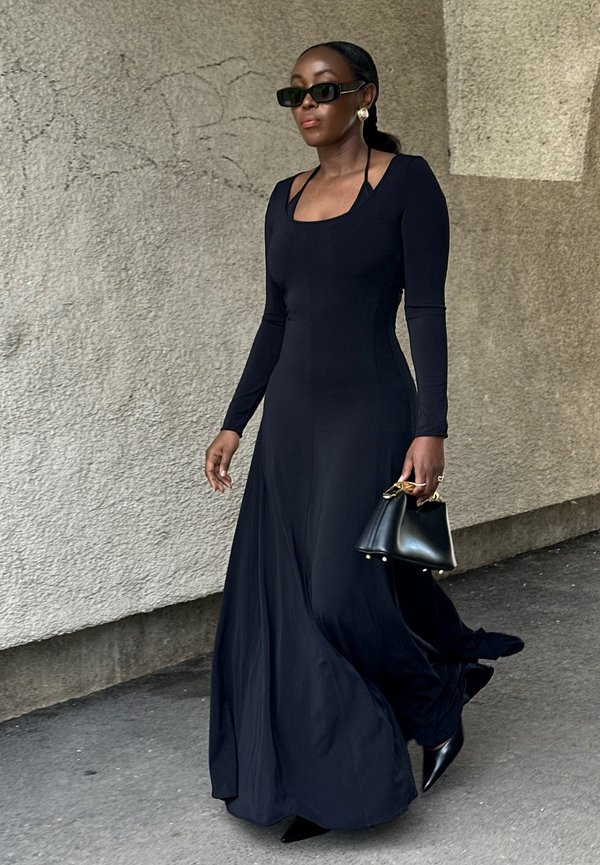 Žena kráča, oblečená v dlhých čiernych šatách a slnečných okuliaroch, nesie čiernu kabelku so zlatými rukoväťami na chodníku.