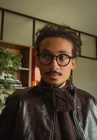 Svart faux skinnjakke med høy krage, brukt over en mørk strukturert topp. Sirkulære briller fremhever ansiktet, rammet inn av krøllete hår.