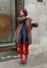 Leopardenmuster-Mantel über einem marineblauen Kleid, rote Strumpfhosen und grüne Plateauschuhe mit roten Akzenten. Der Hintergrund zeigt eine strukturierte Stein- und Metalltür.