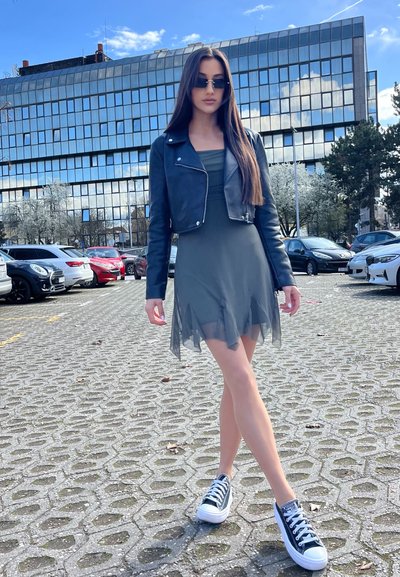 Chaqueta de cuero negra corta, vestido gris ajustado con dobladillo transparente y zapatillas negras y blancas; de pie sobre un pavimento texturizado con coches de fondo.