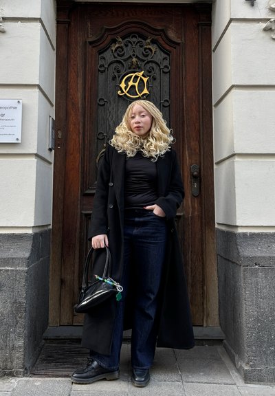 Abrigo largo negro sobre una blusa negra, jeans azules y zapatos negros. Sosteniendo un pequeño bolso negro con un llavero colorido. Puerta de madera al fondo.