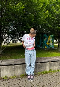 Weißes T-Shirt mit pinkem "NIKE"-Text, hellen, lockeren Jeans, Sneakers mit pinken Akzenten und einem silbernen Gürtel. Stehend auf einer Holzbank.