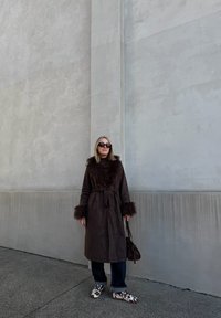 Barna szőrrel díszített kabát hosszú ujjakkal, a deréknál megkötve; sötét farmernadrággal és barna pöttyös mintájú fehér cipővel párosítva.