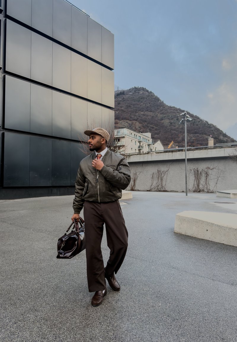 Homme en veste bomber verte et casquette marchant à l'extérieur près d'un bâtiment moderne, portant un sac noir, avec en arrière-plan une montagne et un paysage urbain.