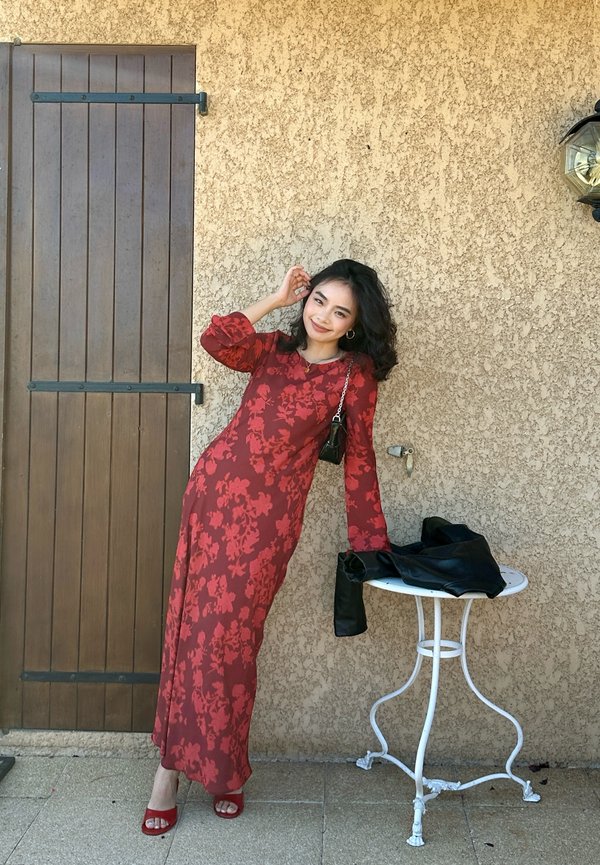 Femme en robe fleurie rouge et talons assortis appuyée contre un mur beige près d'une table ronde blanche avec une veste noire dessus.