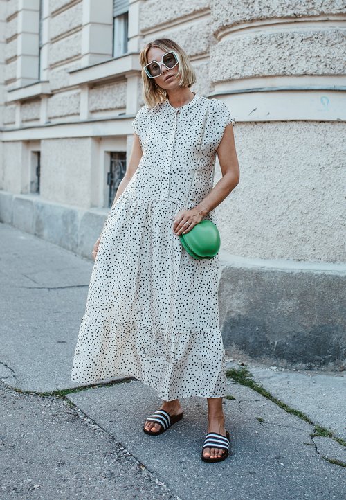 Weißes Kleid mit schwarzen Pünktchen, Knopfverschluss vorne, gestufter Saum. Grünes Handtasche, schwarz-weiße gestreifte Slides. Einheitliche Hintergrundtextur.