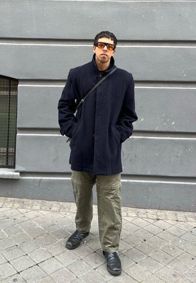 Joven con abrigo oscuro, gafas de sol con tonos naranjas, pantalones oliva y zapatos negros, de pie con las manos en los bolsillos contra una pared gris.