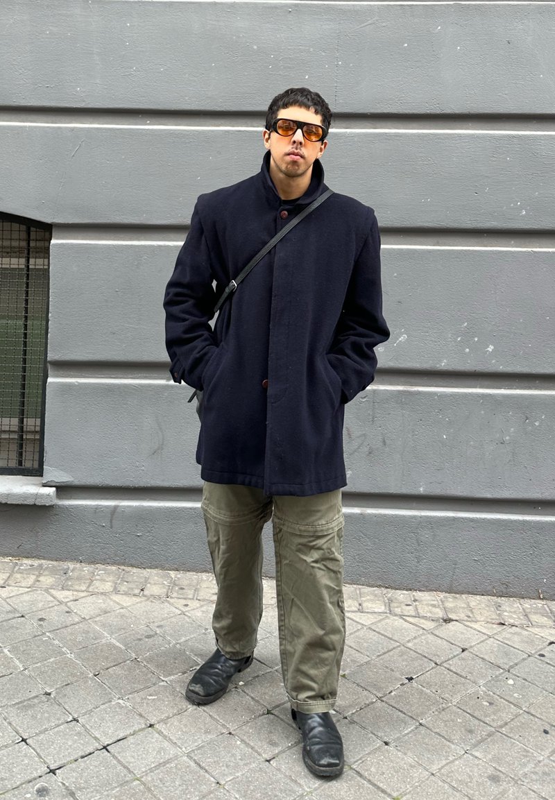 Junger Mann trägt dunklen Mantel, orange getönte Sonnenbrille, olivfarbene Hose und schwarze Schuhe und steht mit den Händen in den Taschen vor einer grauen Wand.