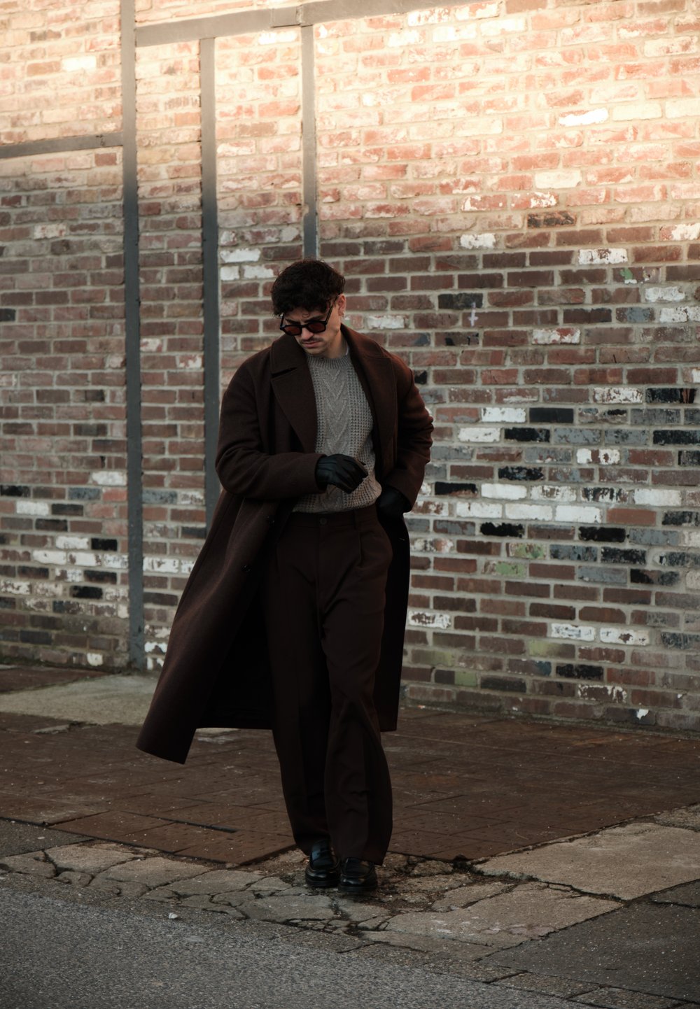 Homme portant un long manteau sombre, un pull gris, des gants noirs et des lunettes de soleil, marchant le long d'un mur en briques sur un trottoir fissuré.
