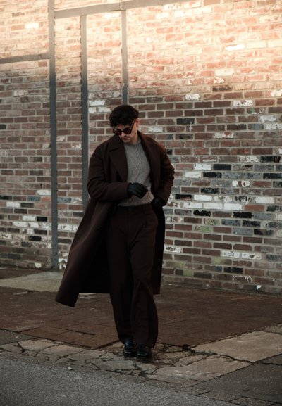 Hombre con abrigo largo oscuro, suéter gris, guantes negros y gafas de sol caminando junto a una pared de ladrillos sobre una acera agrietada.