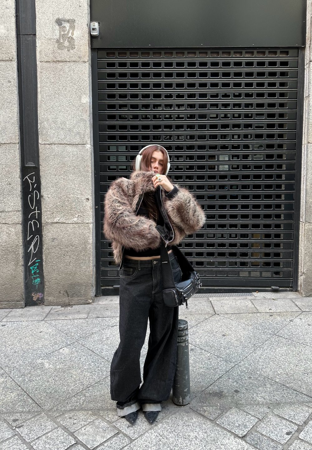 Jeune personne portant une veste grise en tissu duveteux, un jean noir à jambes larges, des chaussures pointues et un casque blanc, debout sur le trottoir en ville près d'un bollard métallique.