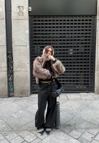 Jeune personne portant une veste grise en tissu duveteux, un jean noir à jambes larges, des chaussures pointues et un casque blanc, debout sur le trottoir en ville près d'un bollard métallique.