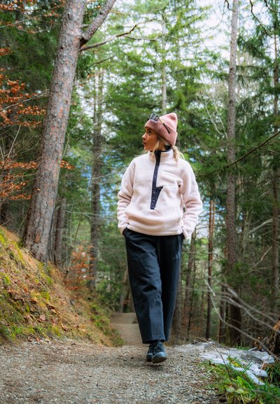 Sudadera de forro polar en rosa claro con un bolsillo con cremallera negra, combinada con pantalones negros y zapatos negros; el modelo lleva un gorro de punto rosa. Fondo de bosque.