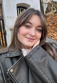 Veste en cuir noire avec des accents de fermetures éclair et de boutons en argent, portée sur un pull blanc. L'arrière-plan présente un mur en pierre et des feuilles tombantes.