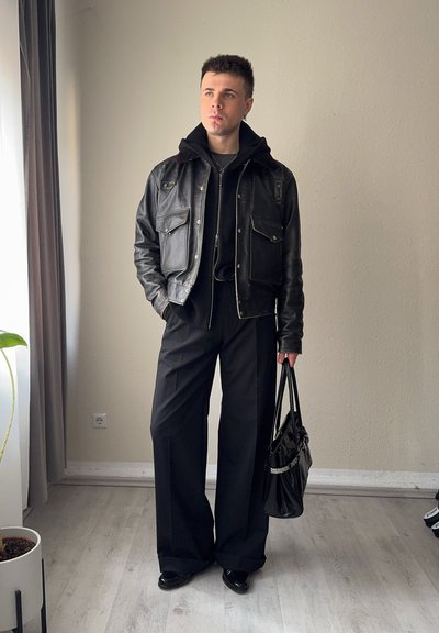 Joven con chaqueta de cuero negra y pantalones de pierna ancha, sosteniendo un bolso negro, de pie en el interior cerca de una ventana con una planta cercana.