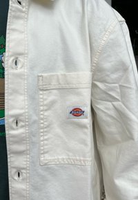 Bela denim srajca z sprednjim žepom, ki ima barvit Dickies znak, vidne šive in podrobnosti z gumbi.