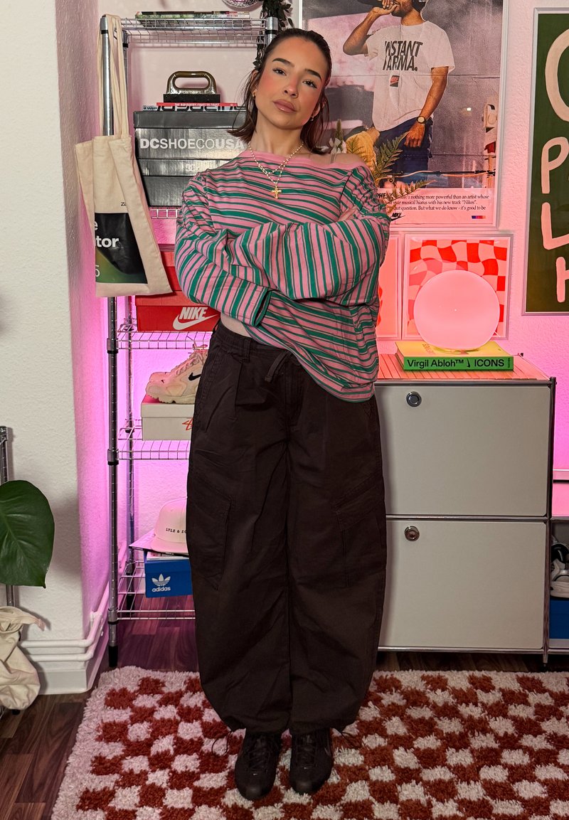 Jeune femme portant une chemise rayée et un pantalon cargo noir debout dans une pièce colorée avec des baskets, des posters et des livres.