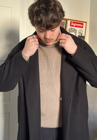 Sort uldfrakke over en beige ribstrikket sweater med høj hals. Person justerer frakkekraven, og sølvringe ses på fingrene.