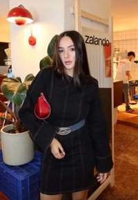 Robe en denim noir à manches longues avec coutures apparentes, accessoirisée d'une pochette rouge et d'une ceinture bleue. Cadre intérieur avec des plantes.