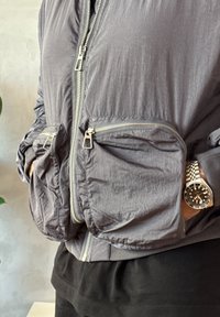 Giacca stile bomber grigia con due tasche frontali con cerniera, tessuto leggero, orlo a costine e una consistenza liscia leggermente arricciata.