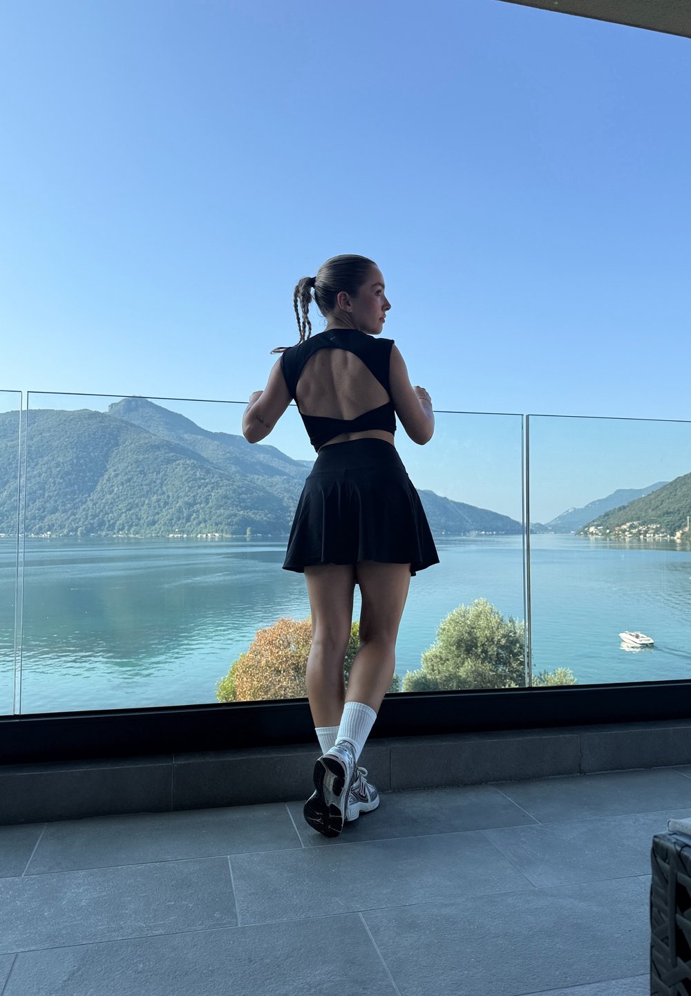 Fekete ruhát és sportcipőt viselő nő egy üvegkorláton támaszkodik, miközben a tóra és a hegyekre néz egy tiszta kék ég alatt.
