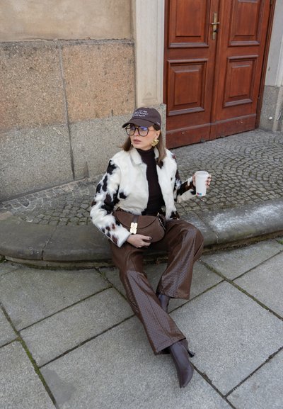 Mujer con chaqueta de piel blanca y negra, pantalones marrones y gorra, sentada en la acera sosteniendo una taza, cerca de una puerta de madera y una pared de piedra.