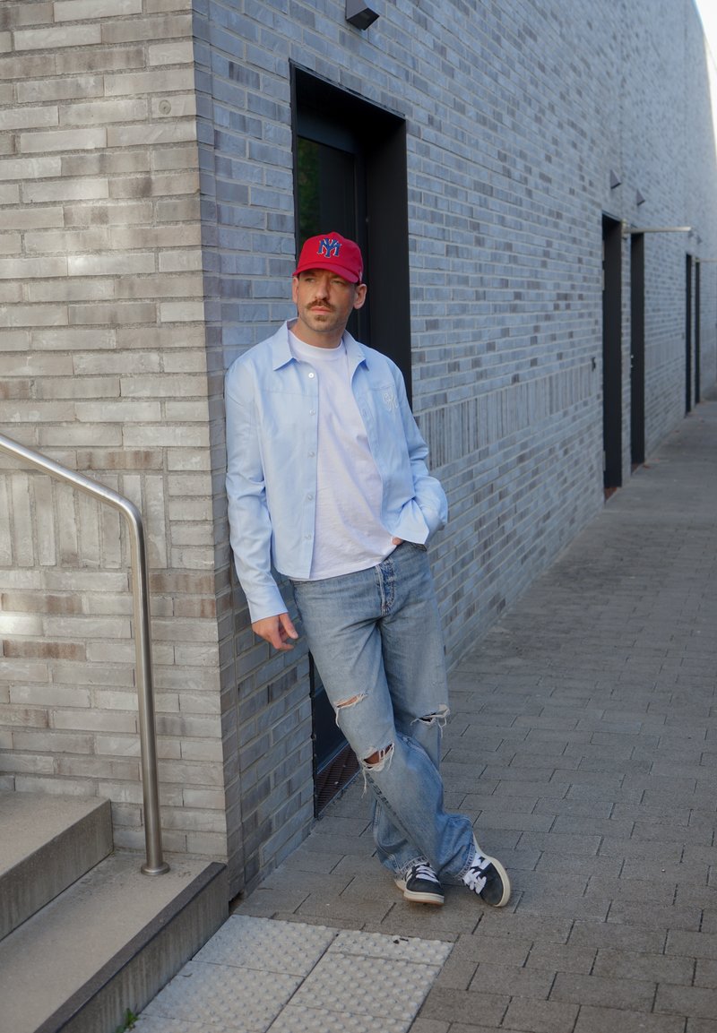 Un homme portant une casquette rouge, une chemise bleu clair, un jean déchiré et des baskets s’appuie contre un mur de briques, à côté d’une porte.