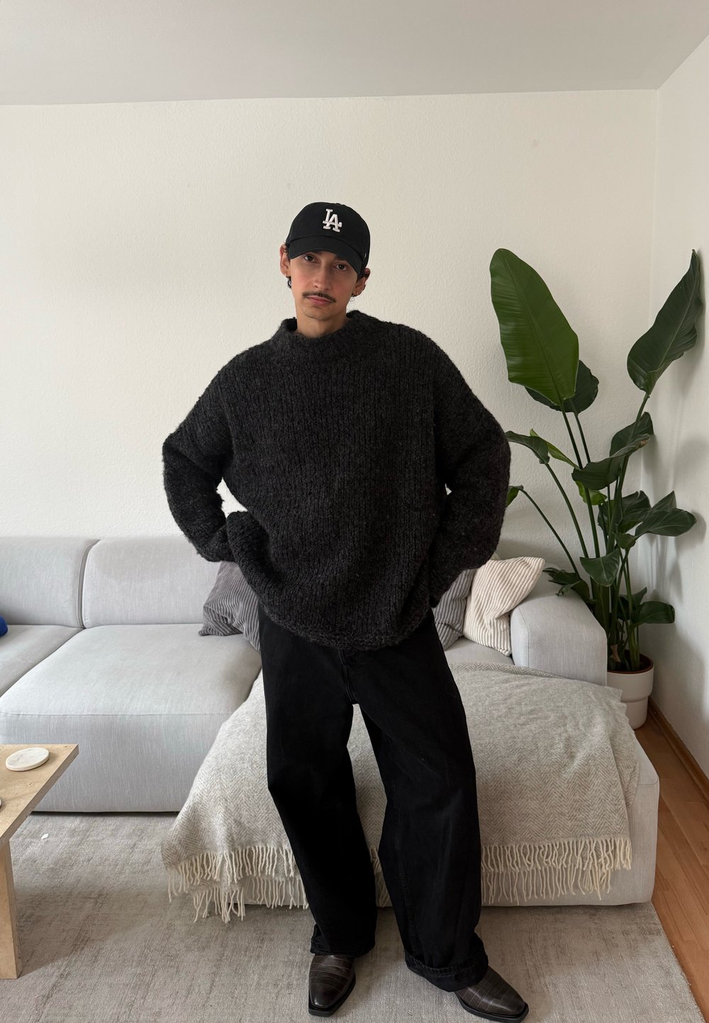Pull en maille épaisse et sombre avec un col montant, pantalon noir à jambes larges, et chaussures en cuir noir. En arrière-plan, une plante et un canapé clair.